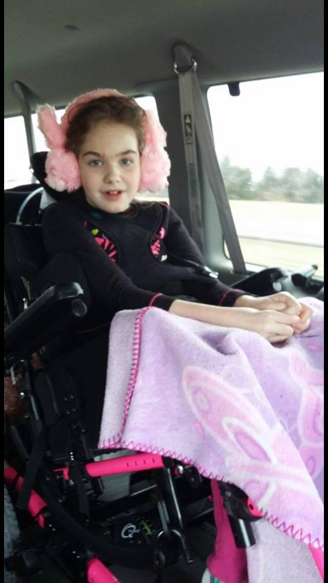 Photo: Abby, wearing pink ear muffs with a pink blanket over her lap, rides in wheelchair-accessible van that was donated to her family.
